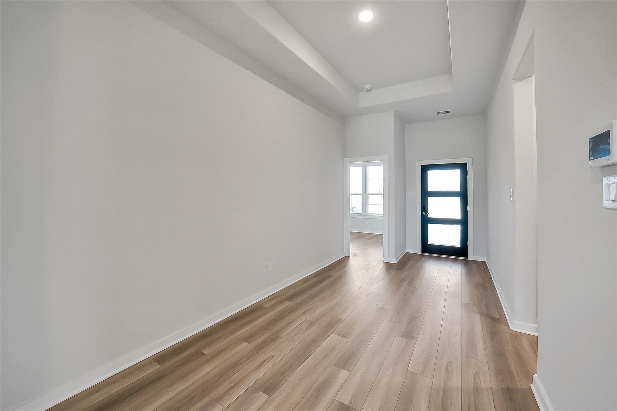 Spacious hallway with tray ceiling, light wood floors, and navy glass door in Davidson Homes Acadia A, Sunterra, Katy, Texas