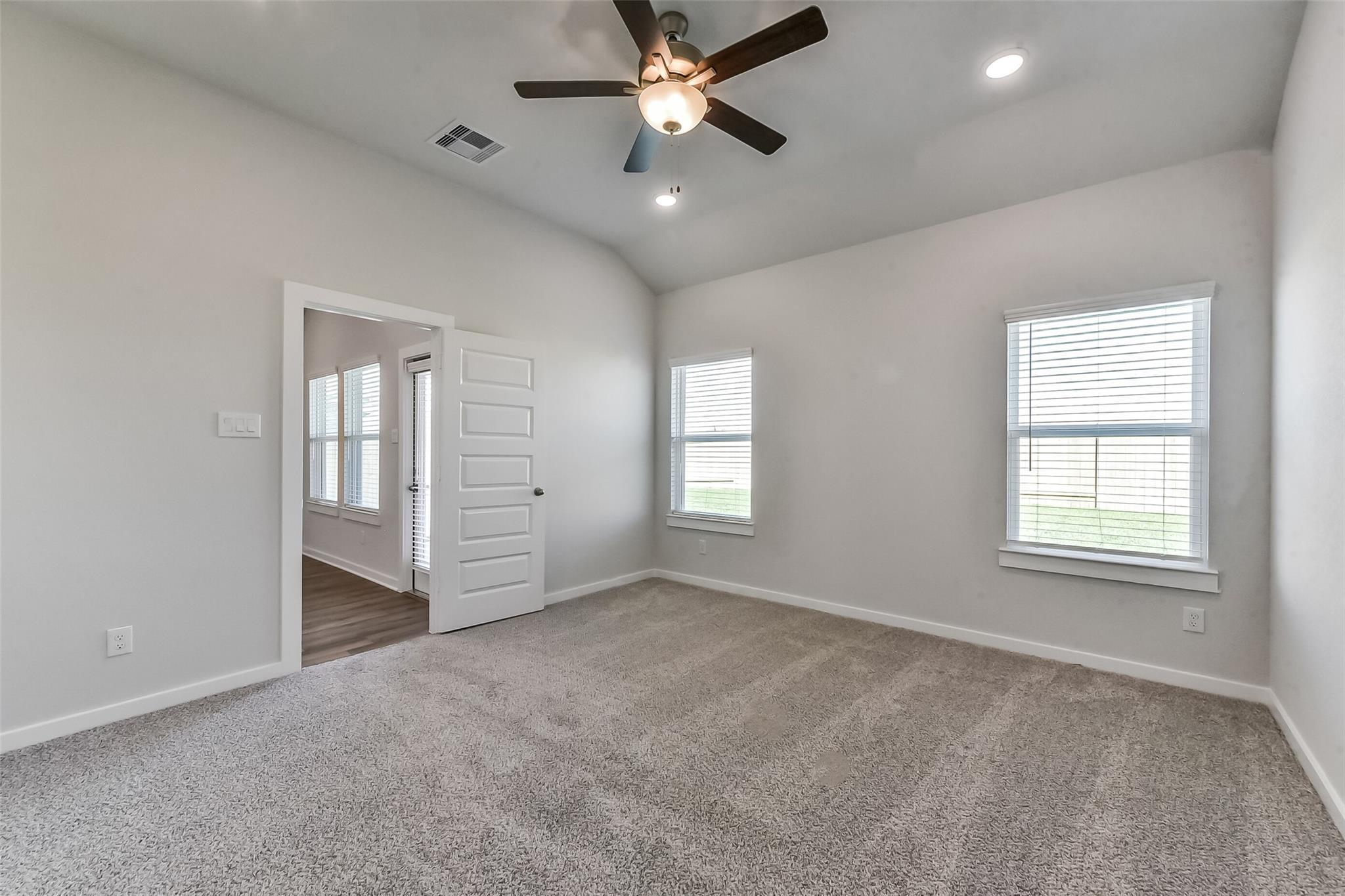 Spacious bedroom with gray carpet, ceiling fan, blinds-covered windows, and open door in Davidson Homes The Costa C, Dayton, Texas