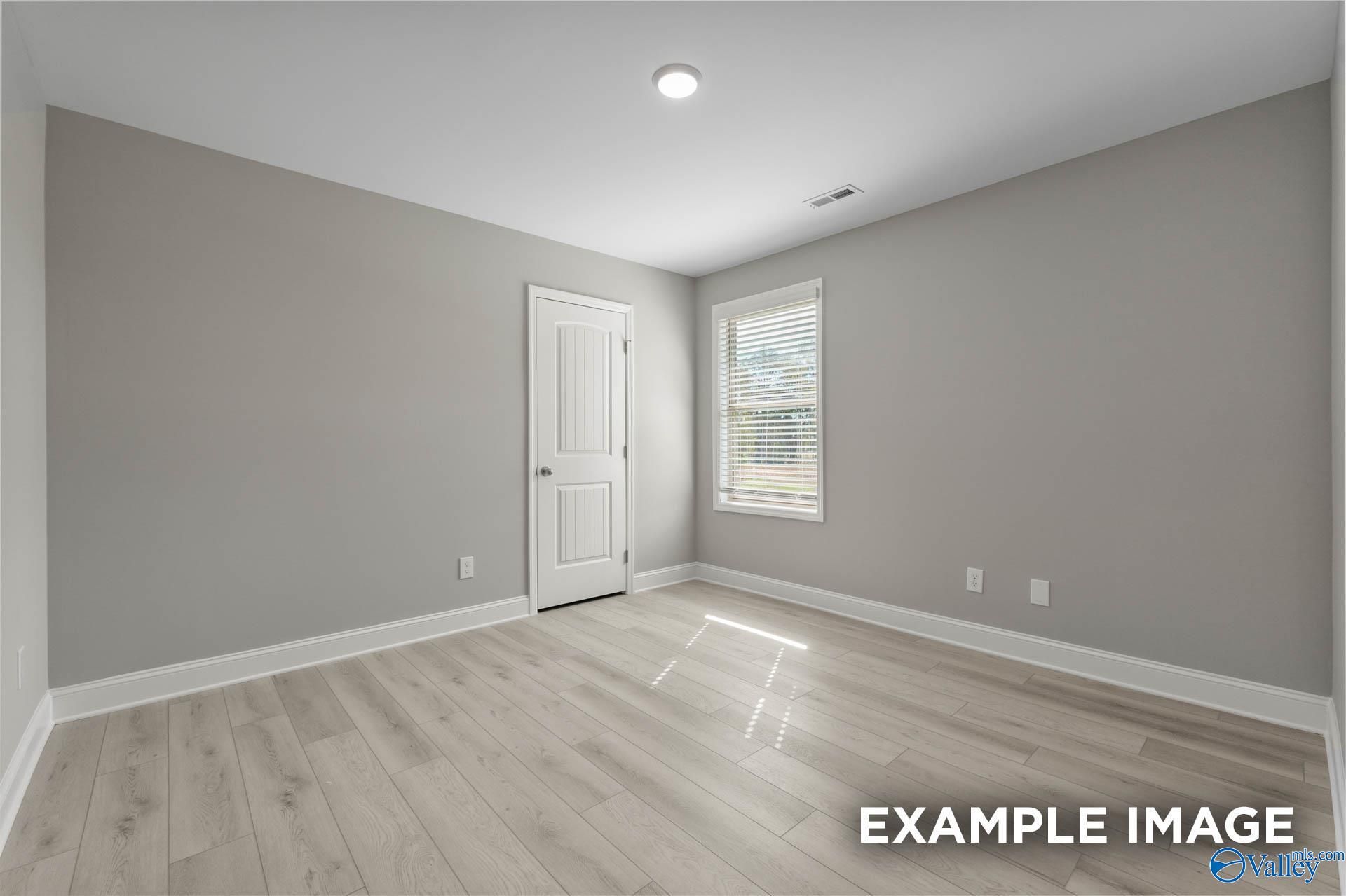 Empty secondary bedroom with light gray walls, white paneled door, blinds-covered window, and oak laminate floors in The Everett B, Toney, Alabama