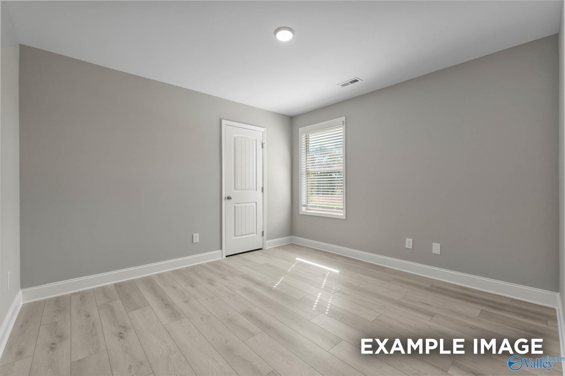 Empty secondary bedroom with light gray walls, white paneled door, blinds-covered window, and oak laminate floors in The Everett B, Toney, Alabama