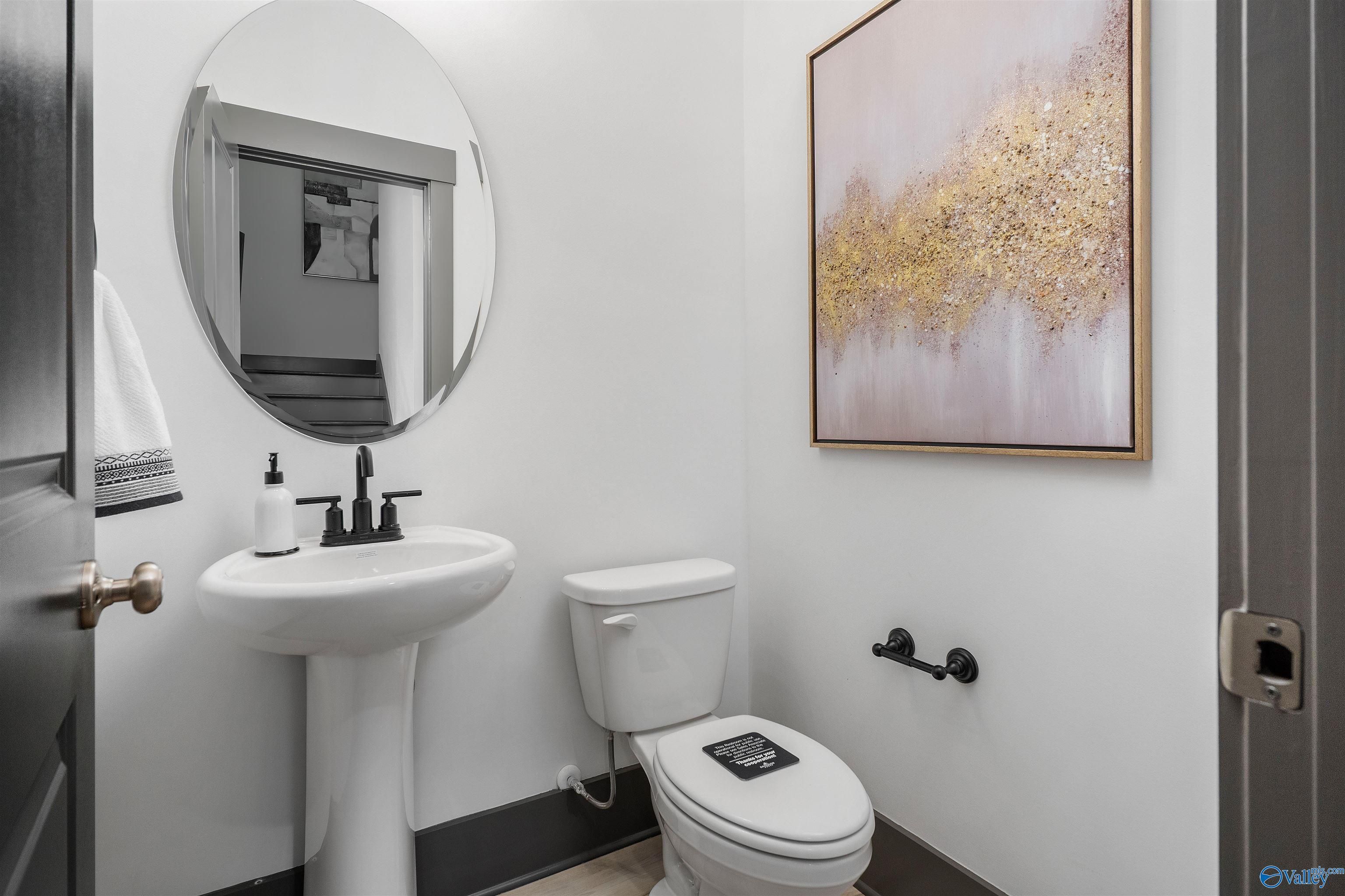 Modern powder room with white pedestal sink, round mirror, and gold abstract art in Davidson Homes The Rockford B, Madison, Alabama