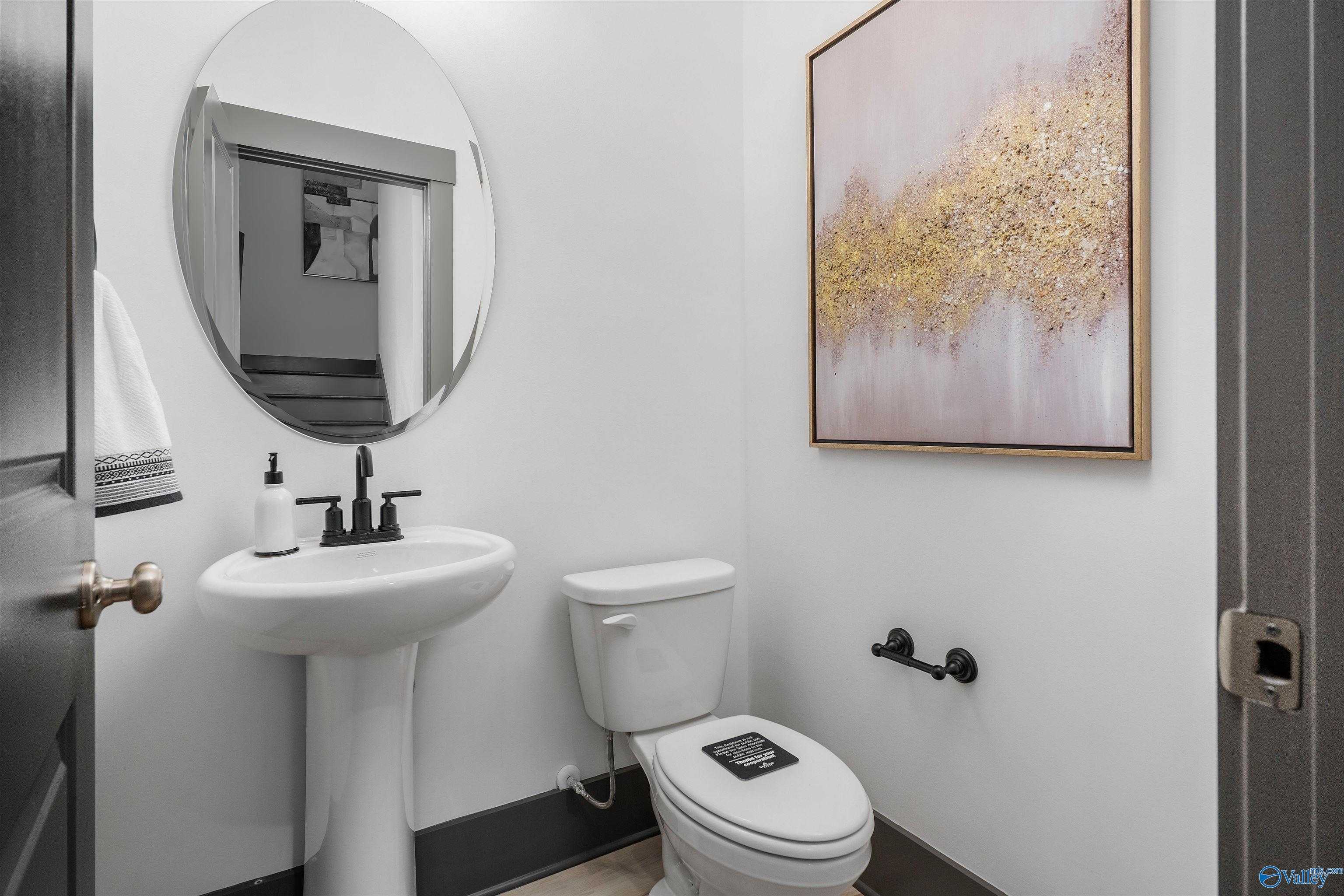 Modern powder room with white pedestal sink, round mirror, and gold abstract art in Davidson Homes The Rockford B, Madison, Alabama