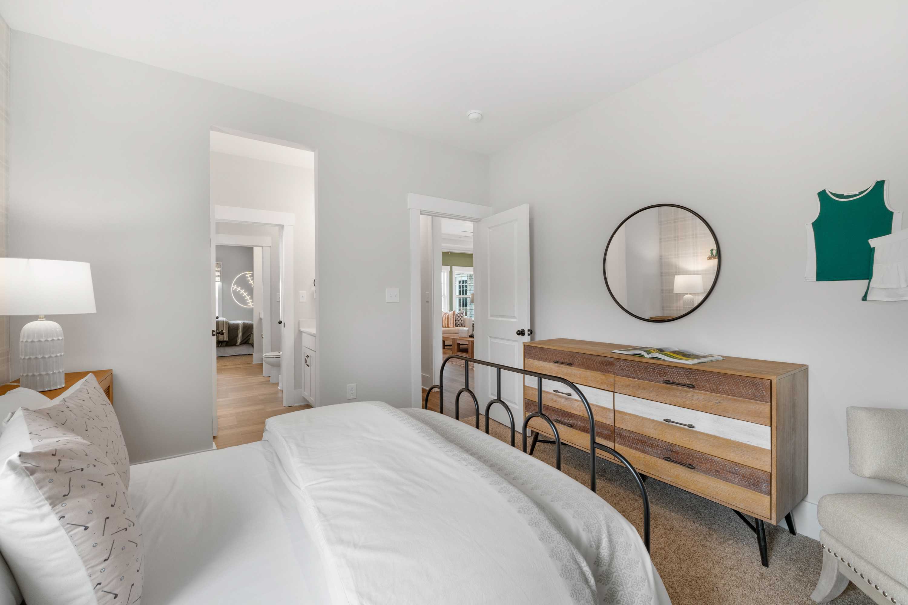 Serene bedroom interior at Briercreek in Meridianville AL featuring white bedding, wooden dresser, round mirror, and open hallway