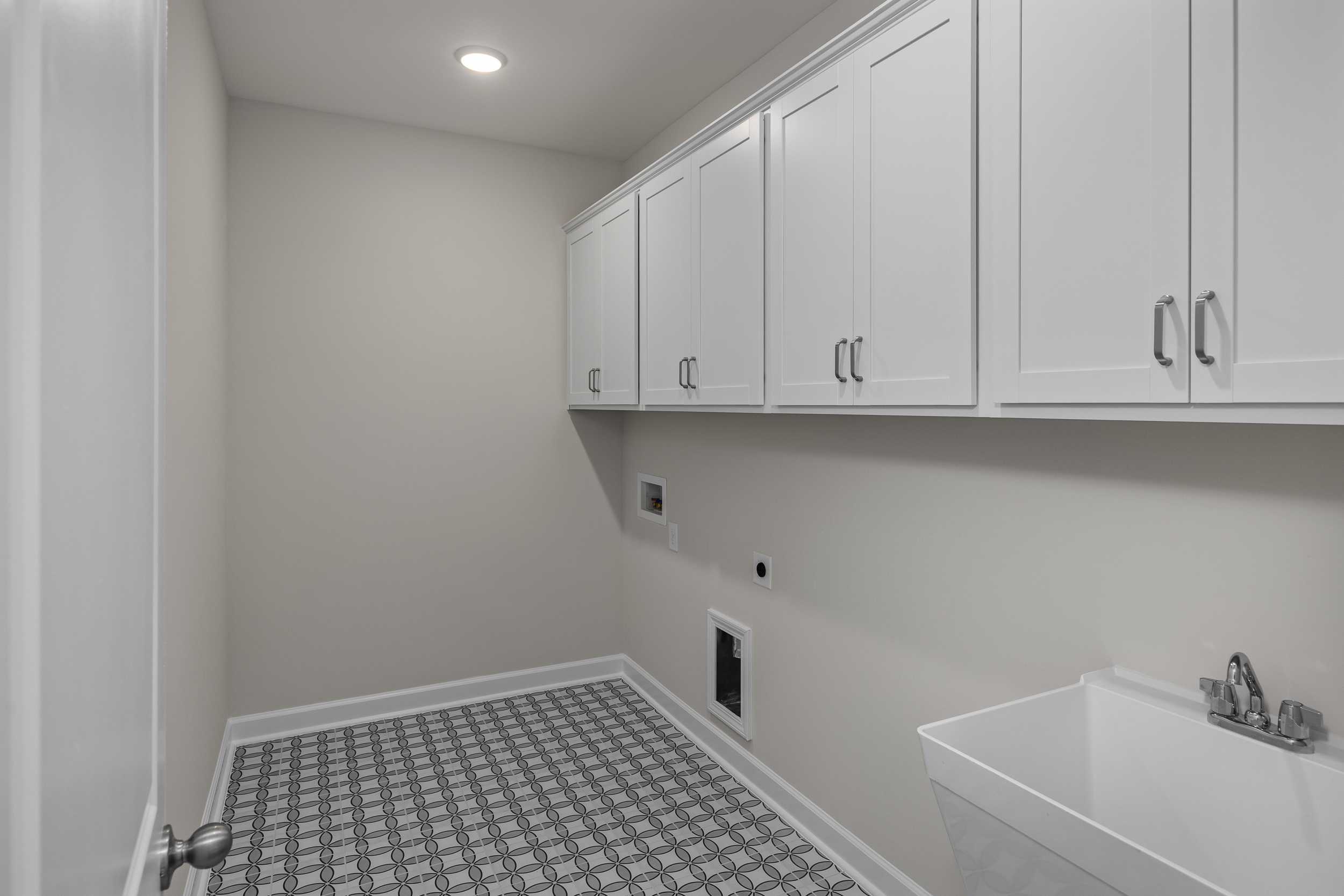 Spacious laundry room in The Aspen by Davidson Homes with white upper cabinets, utility sink, washer-dryer hookups, and gray checkered tile floor