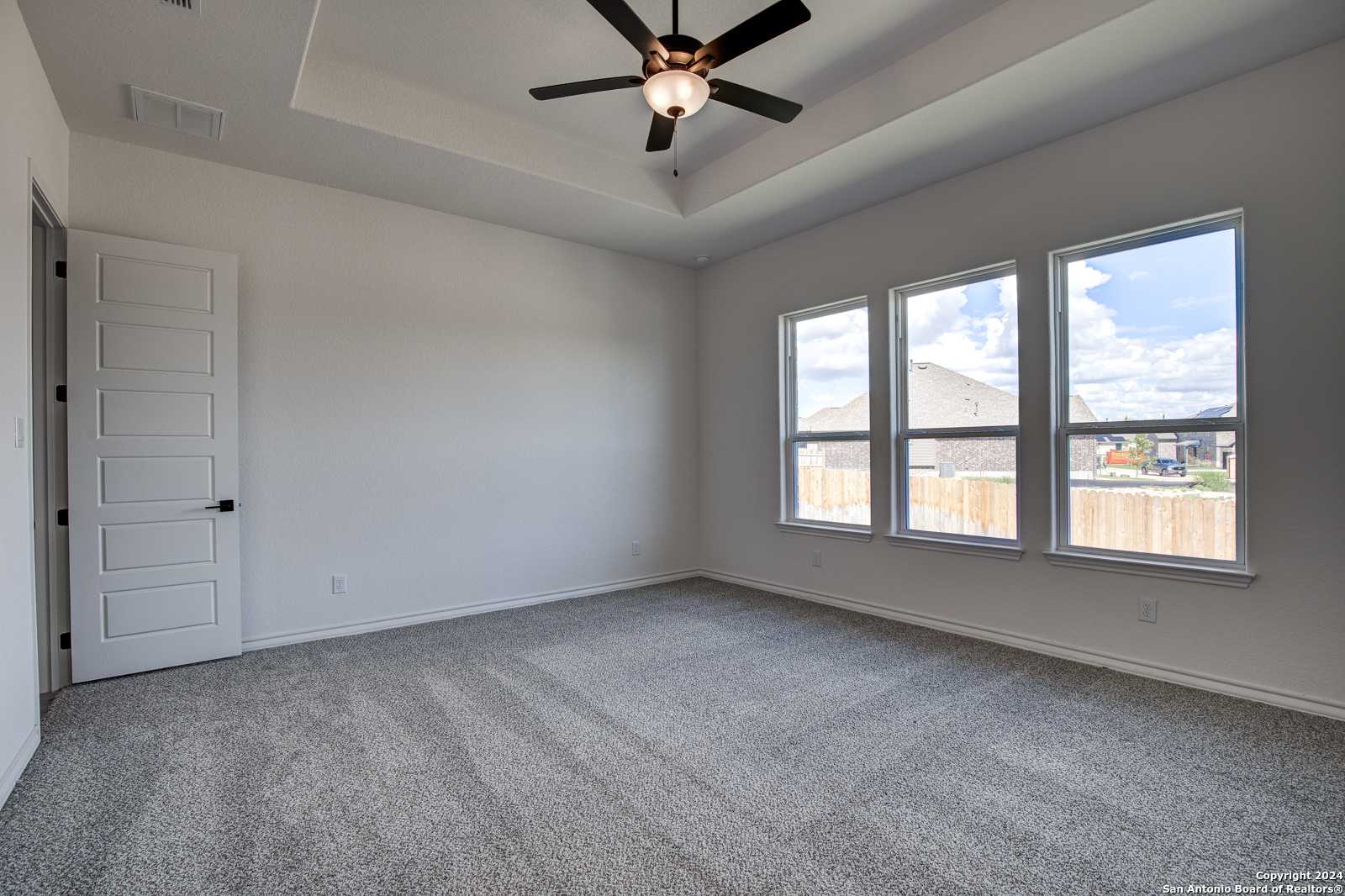 Bright bedroom featuring tray ceiling, ceiling fan, and triple windows with backyard view in Davidson Homes The Garner B, Castroville, Texas