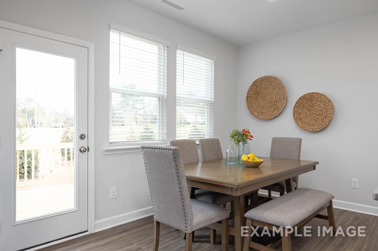Spacious dining area in The Grace A home with wooden table, tufted chairs, bench seating, floral vase, fruit bowl, woven wall decor, large windows, and sliding door to deck