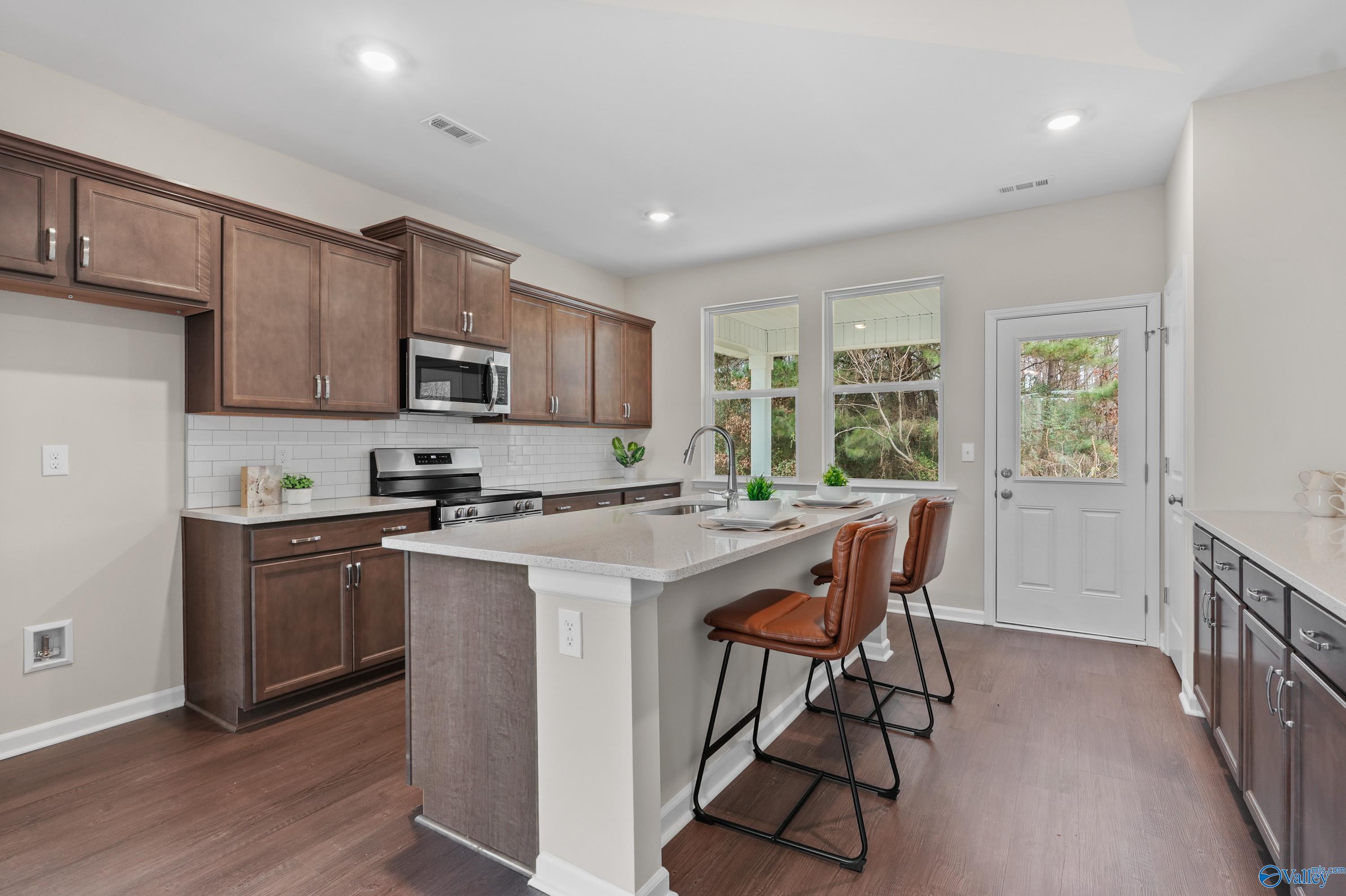 Modern kitchen featuring wood cabinets, stainless steel appliances, quartz island with bar stools, and large windows in The Stella by Davidson Homes, Hazel Green, Alabama