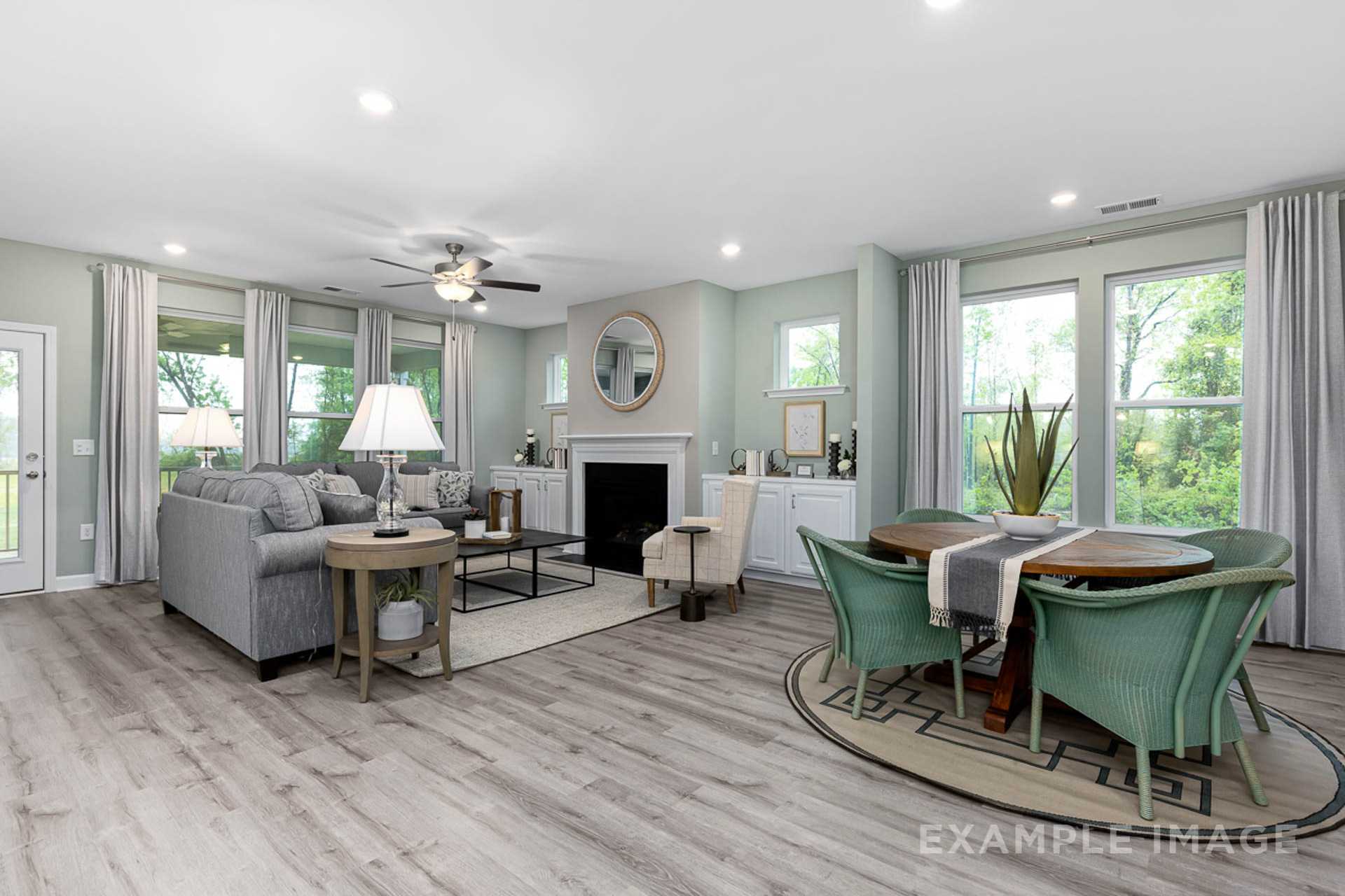 Open-concept living and dining room in Prince Place, Fuquay-Varina NC featuring hardwood floors, gray sofa, green chairs, fireplace, and large windows