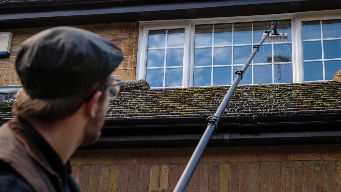 A professional cleaning white uPVC soffits and fascias from the ground, with the roofline becoming visibly brighter against pale brickwork