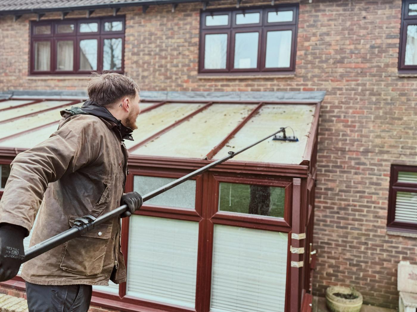 A garden conservatory with a cloudy roof before cleaning, showing leaf debris, algae along the frame lines and soft planting around the structure