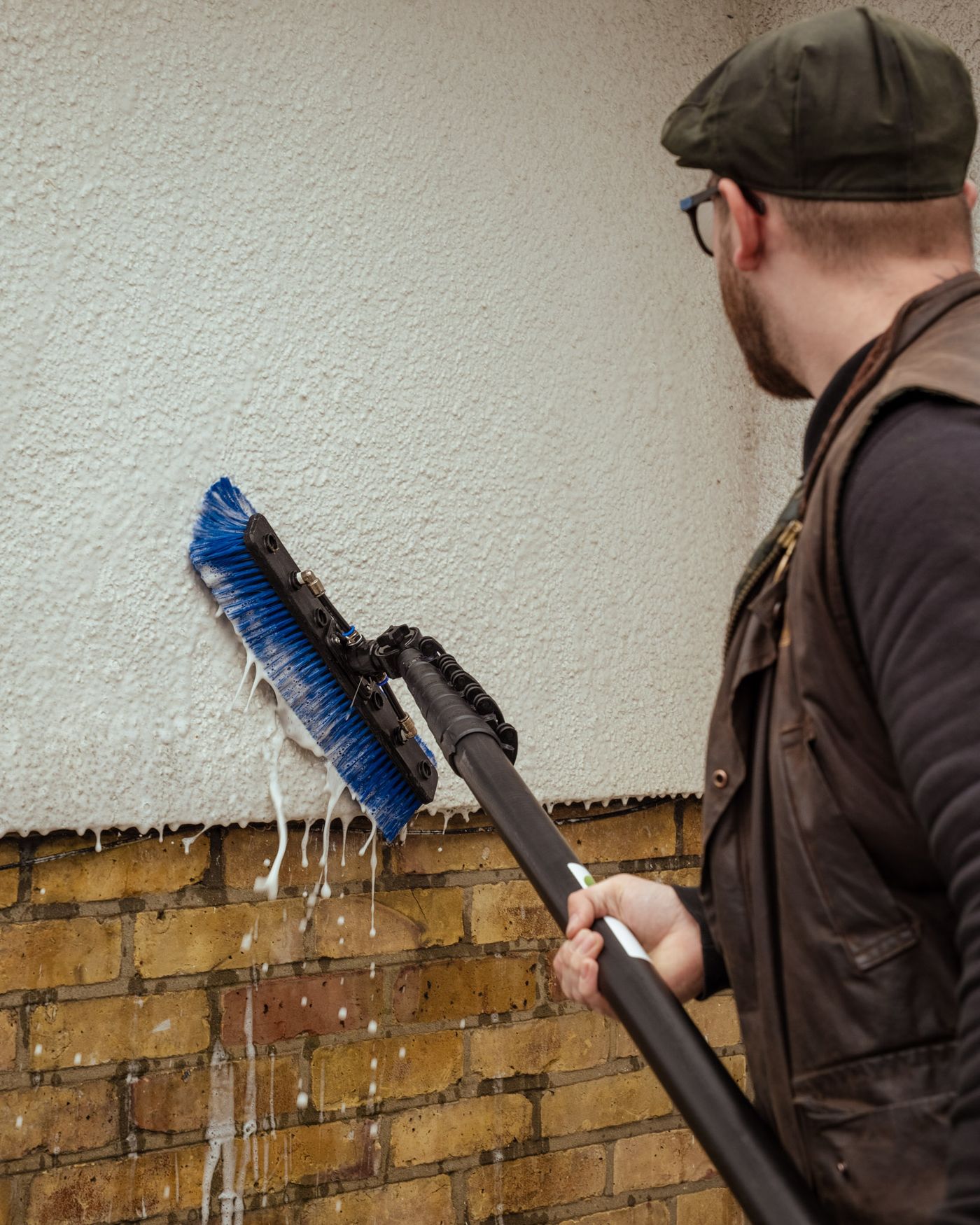 A low-pressure soft washing treatment being applied to a shaded painted masonry wall on a period home, with planting protected and windows closed