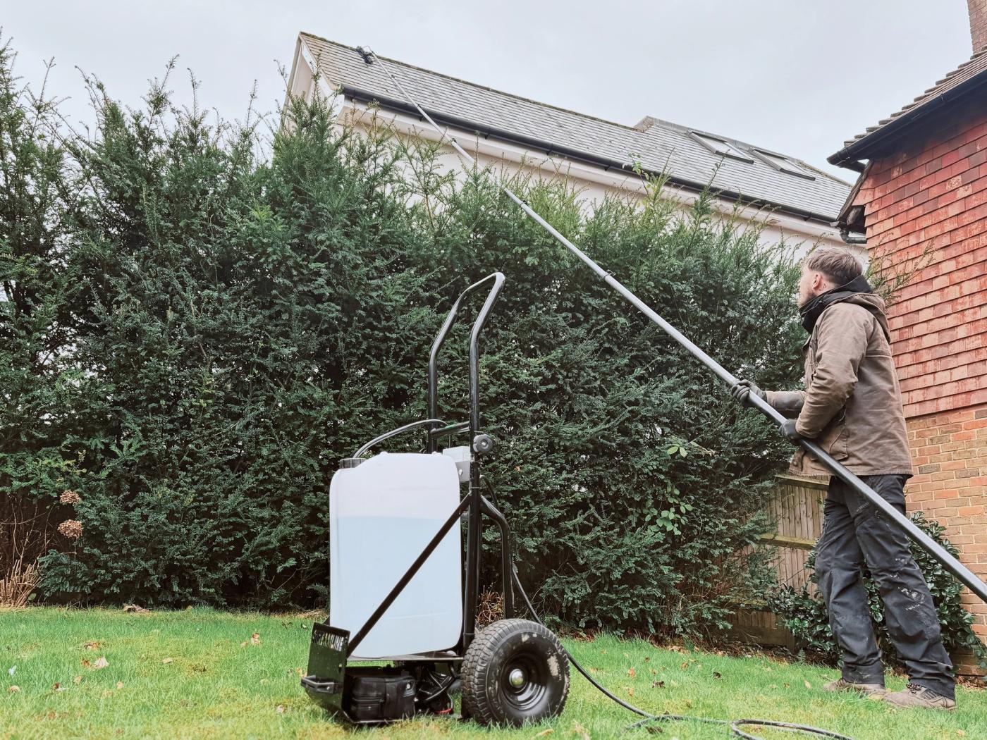 A roof cleaning treatment being applied carefully at low pressure to a tiled roof, with gutters protected and garden planting visible below
