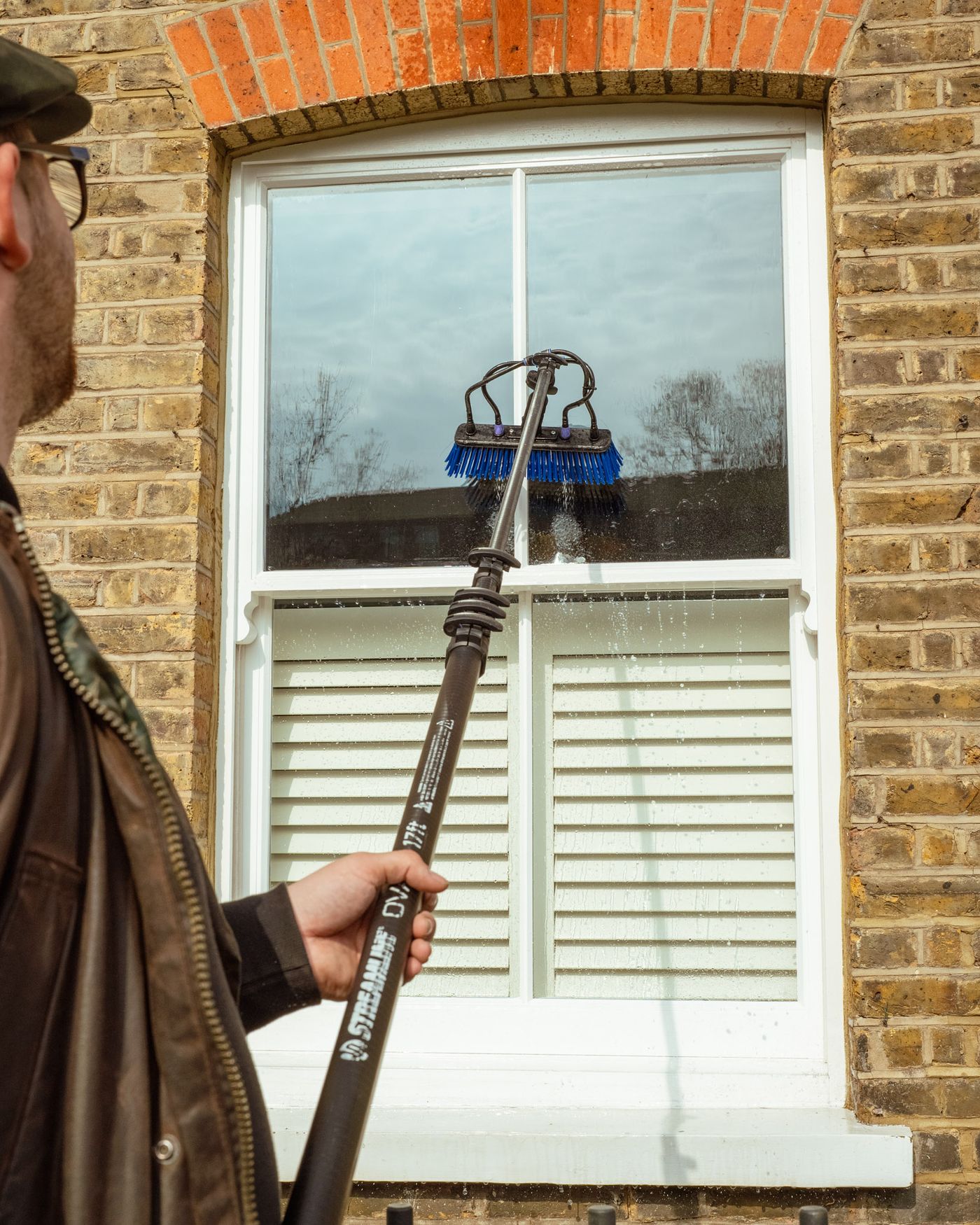 A pure water pole brush rinsing upper-storey sash windows from the ground, with water sheeting cleanly down the glass and no ladders against the property