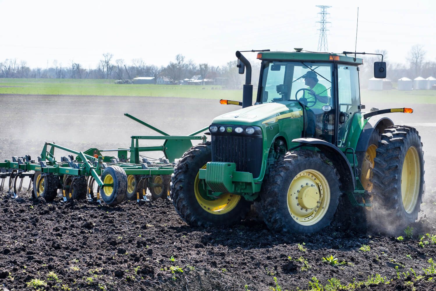 Farmer in tractor tilling farm field preparing soil for spring corn planting. John Deere, power lines