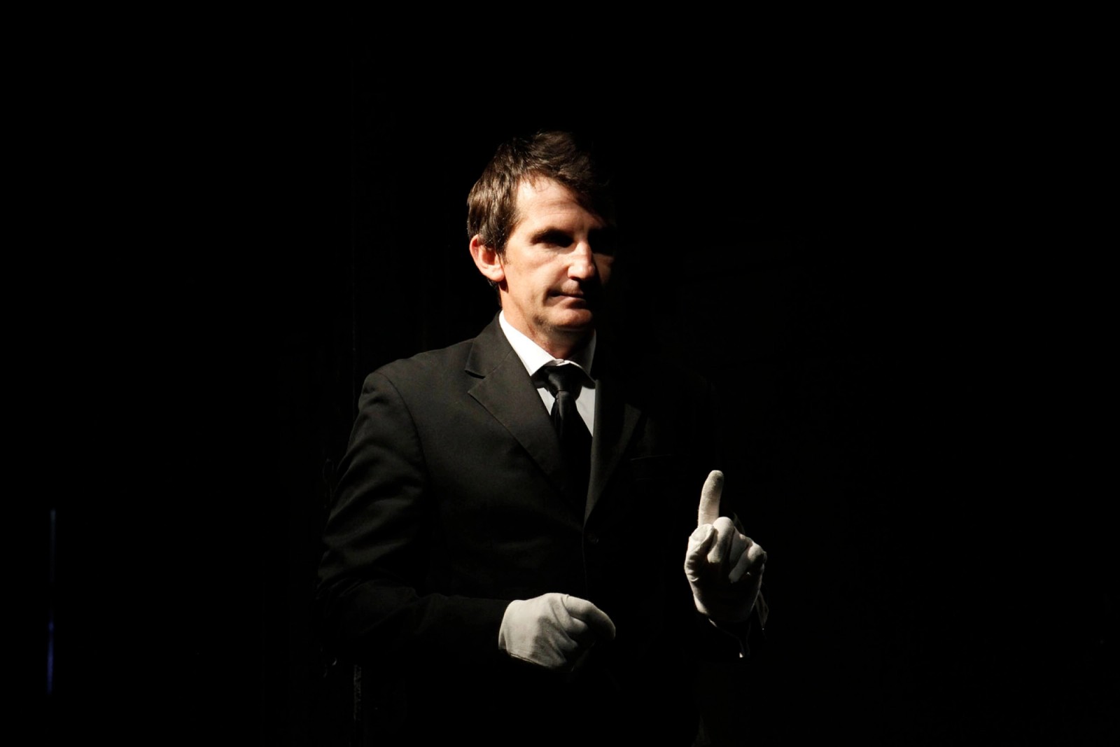 A man wearing a suit and white gloves in a black space as part of Captives of the City by Chamber Made
