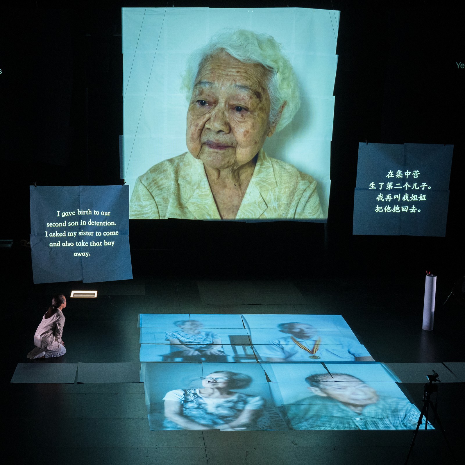 Woman kneeling on stage with multiple large projections of photographic portraits of elderly people.