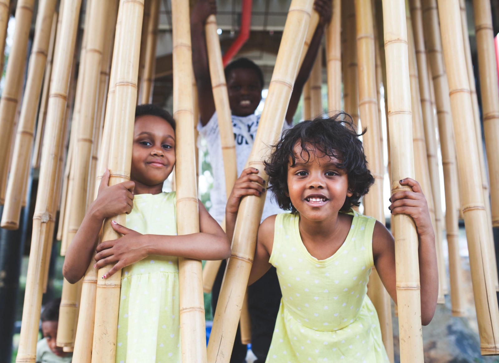 Three children peer cheekily through a bamboo installation as part of The Chamber Made Venny Celebration 2018