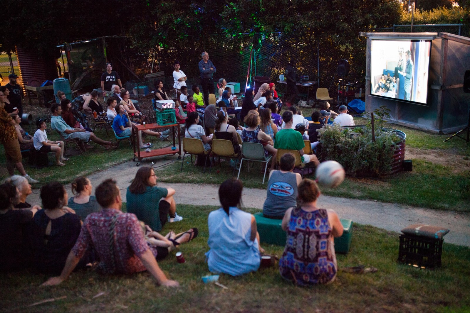 A crowd of people in evening light gather to watch an outdoor screen as part of The Chamber Made Venny Celebration 2018
