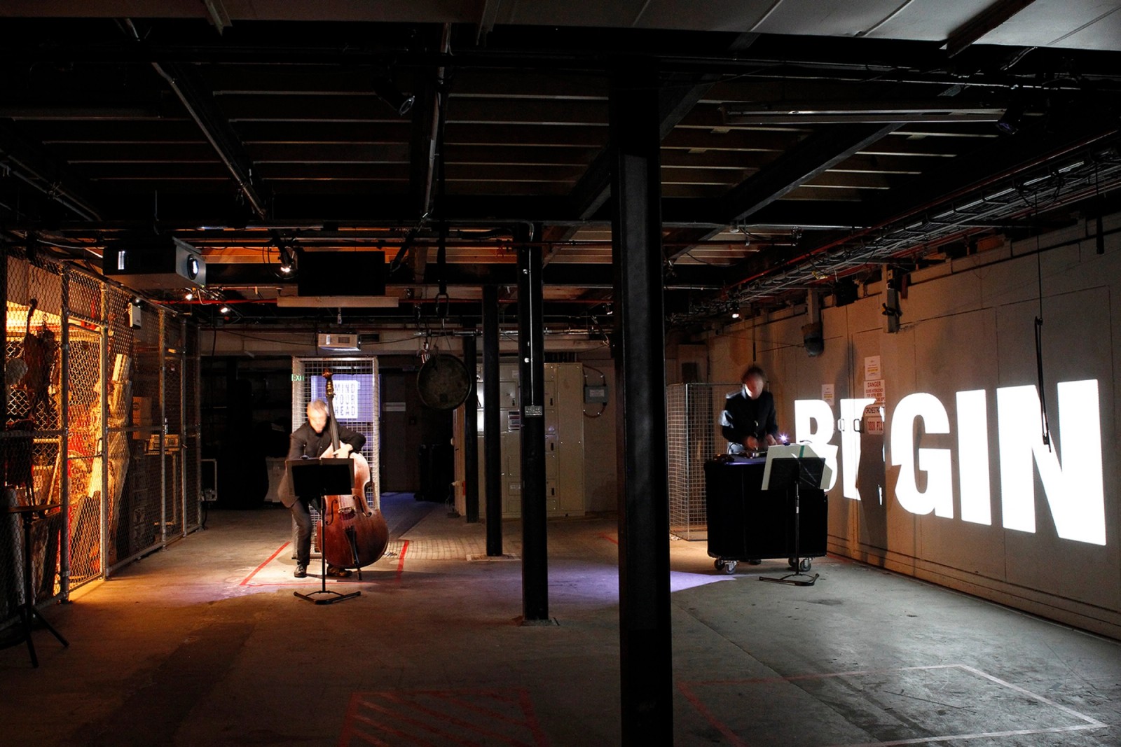 A man with a double bass and one at a small desk in a basement with equipment and a large projection of the word BEGIN as part of Captives of the City by Chamber Made
