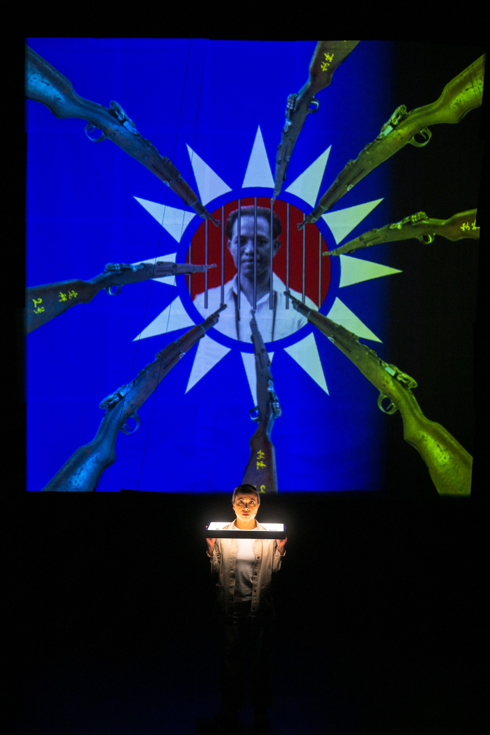 Woman standing on stage in front of large screen with projections of a man's head with many guns aimed towards it as part of One Day We'll Understand.