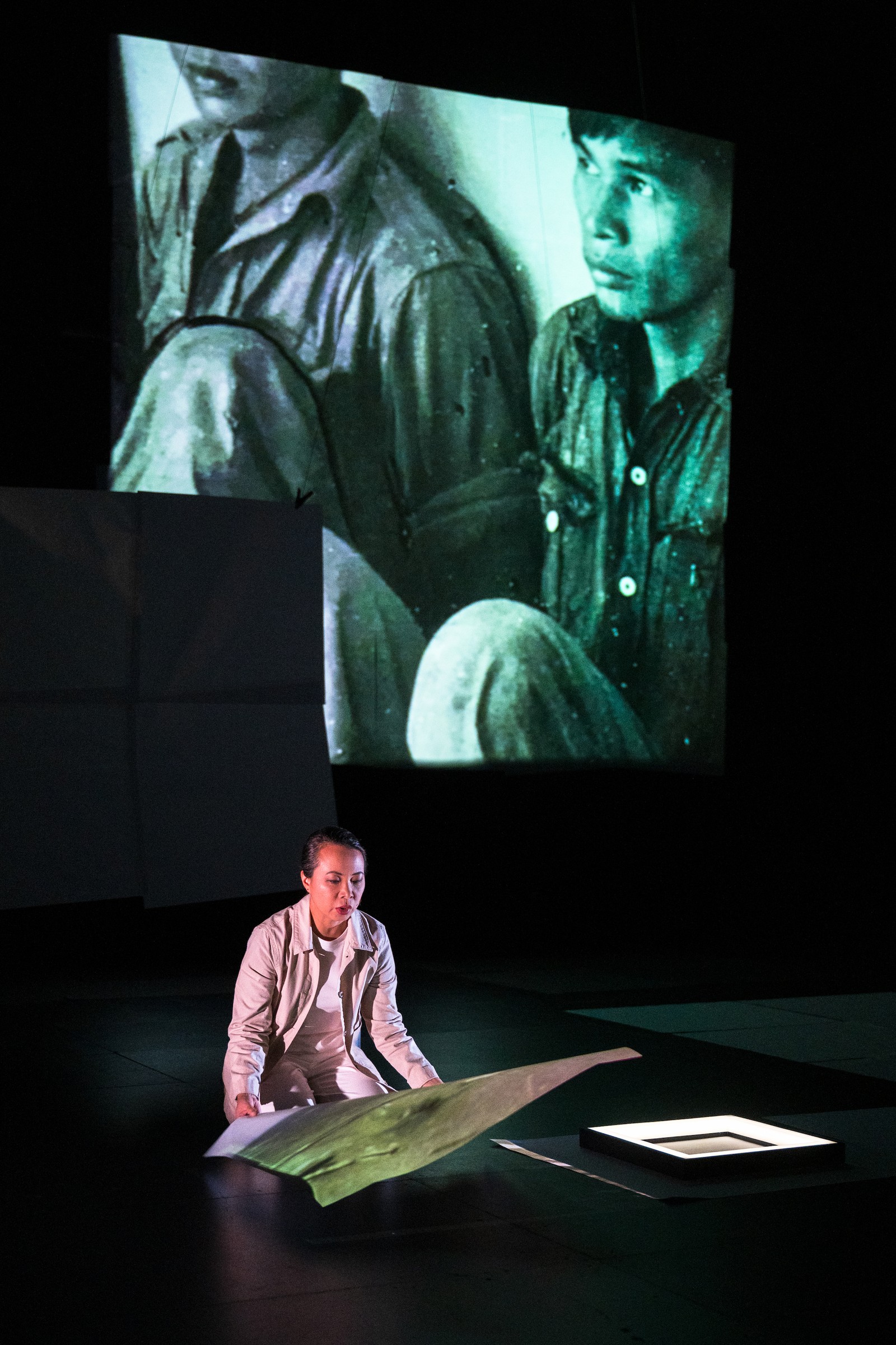 Image of a woman kneeling on stage with a projection of two men on a large screen behind her as part of One Day We'll Understand.
