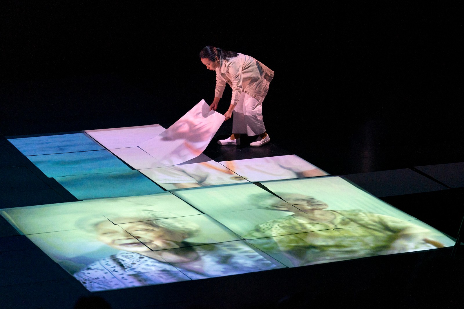 Woman laying out paper on the stage with projected images of portraits of elderly people as part of One Day We'll Understand