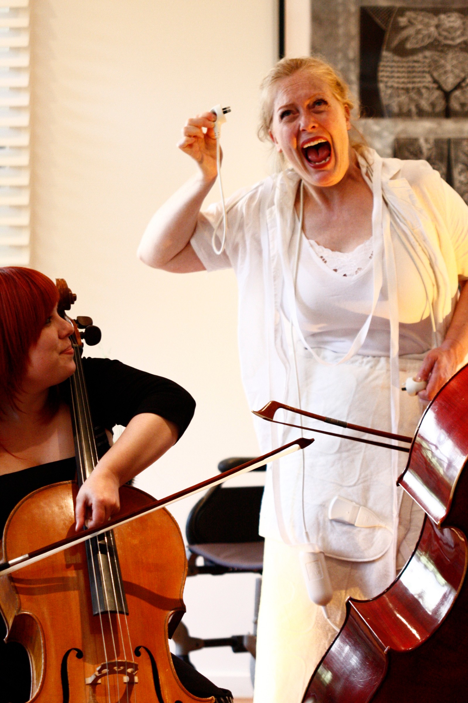 A woman in white with open mouth, with a cellist and double bass nearby performing