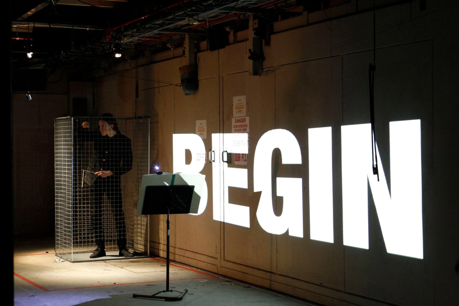 A man in a cage near a wall with the word BEGIN projected onto it as part of Captives of the City by Chamber Made