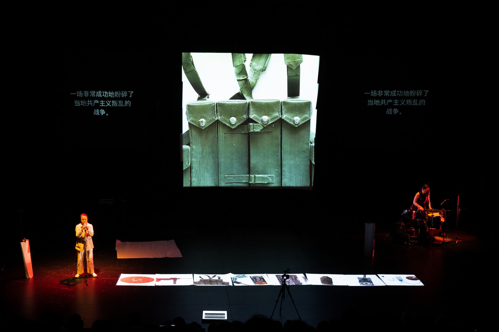 Two performers on stage with a large central projection behind them. One performers is behind a microphone, the other playing a drum kit.