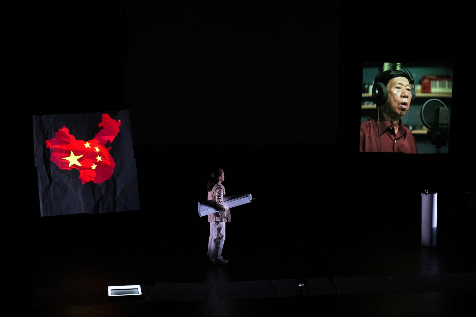A woman on a dark stage standing between two hanging screens. One screen has the image of a Chinese flag  and the other an image of a man singing into a microphone.