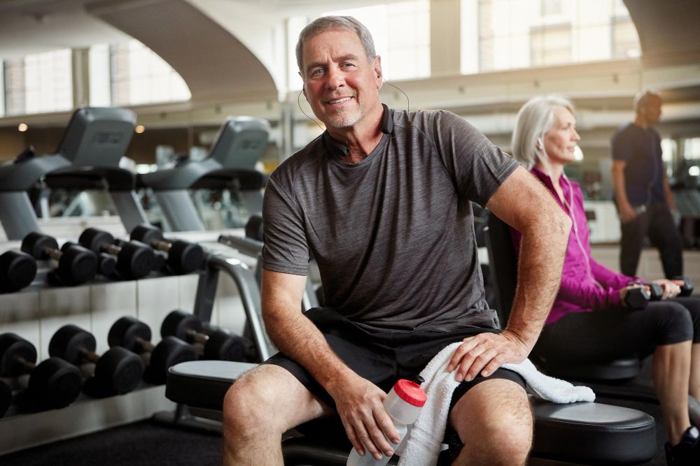 mosolygó férfi mozgás közben leül pihenni az edzőteremben egy padra, kezében törölköző és kulacs, a háttérben hölgy ül