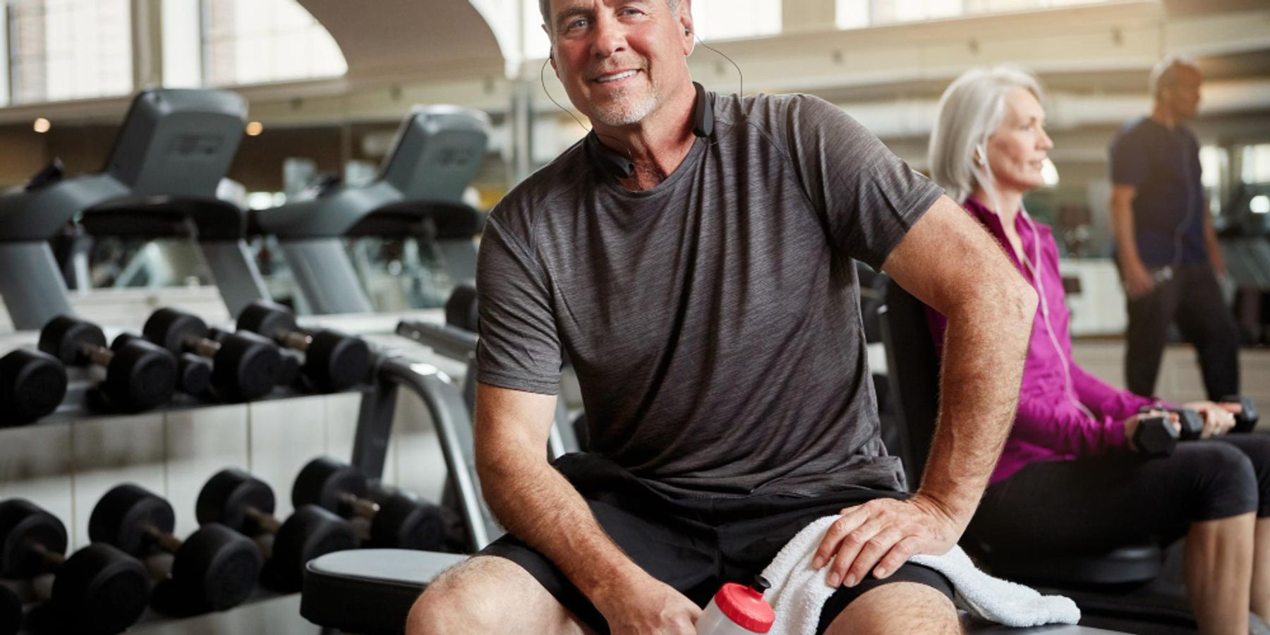 mosolygó férfi mozgás közben leül pihenni az edzőteremben egy padra, kezében törölköző és kulacs, a háttérben hölgy ül