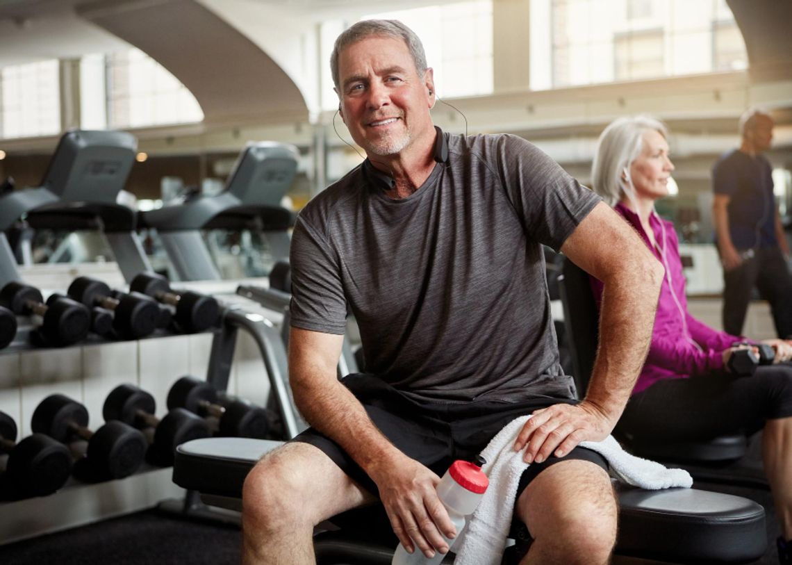 mosolygó férfi mozgás közben leül pihenni az edzőteremben egy padra, kezében törölköző és kulacs, a háttérben hölgy ül