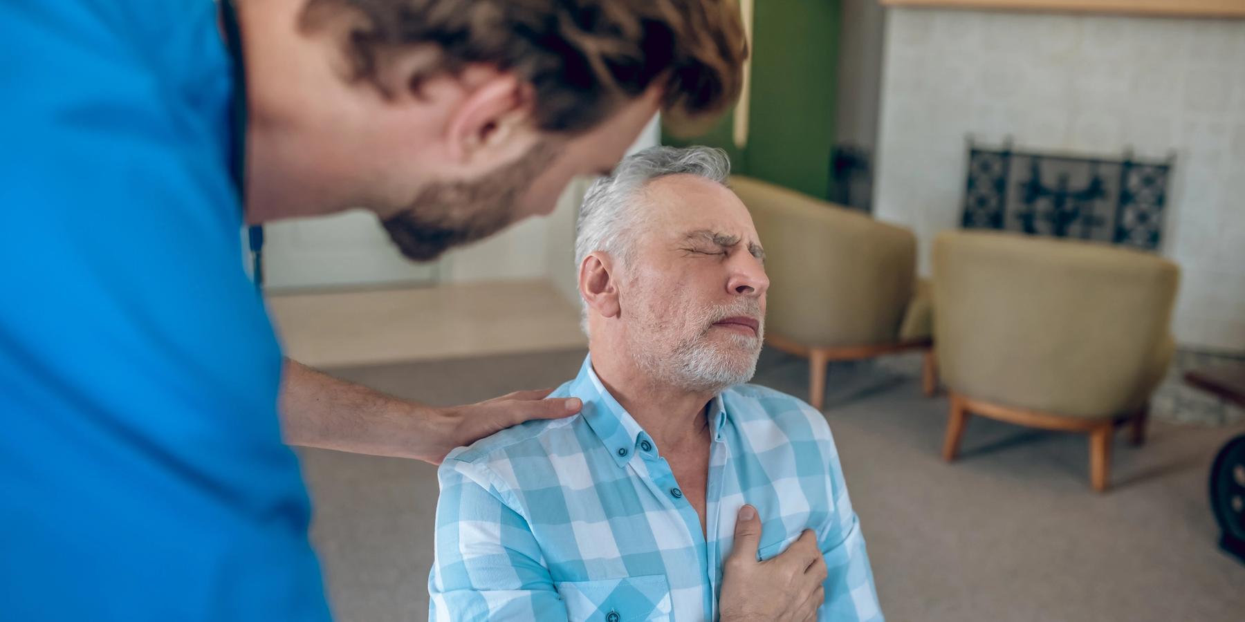 Sötét hajú orvos hajol egy idősödő, kaukázusi férfi felé, aki mellkasi fájdalmat érez.
