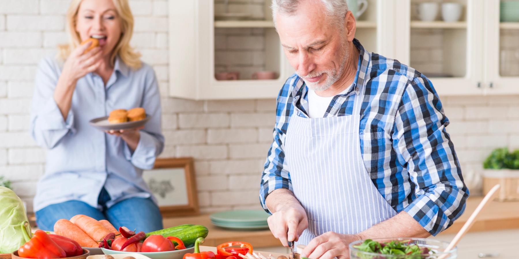 Idősebb férfi vágja a zöldségeket a vágódeszkán, miközben a felesége a háttérben a konyhában muffint eszik.