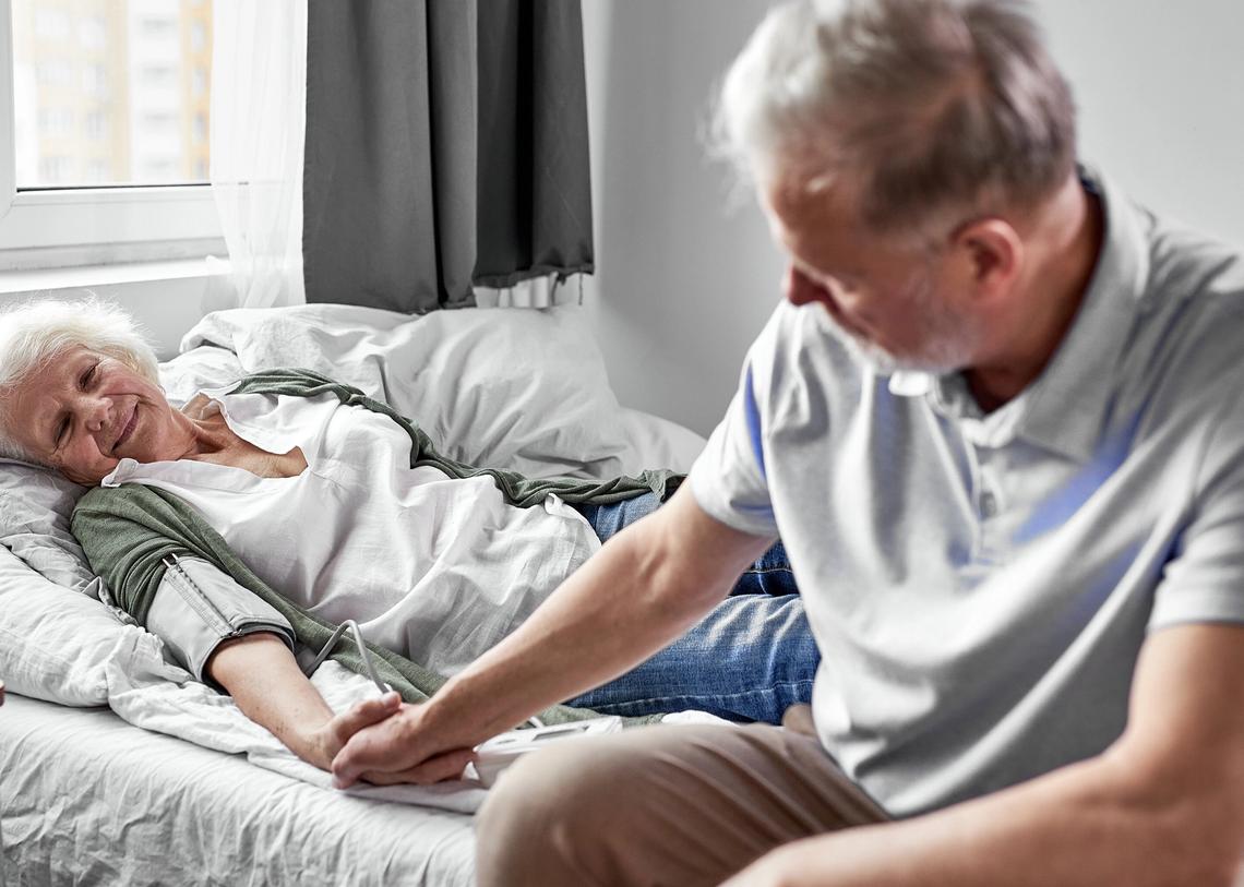idős férfi fogja az ágyban fekvő beteg felesége kezét