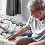 idős férfi fogja az ágyban fekvő beteg felesége kezét