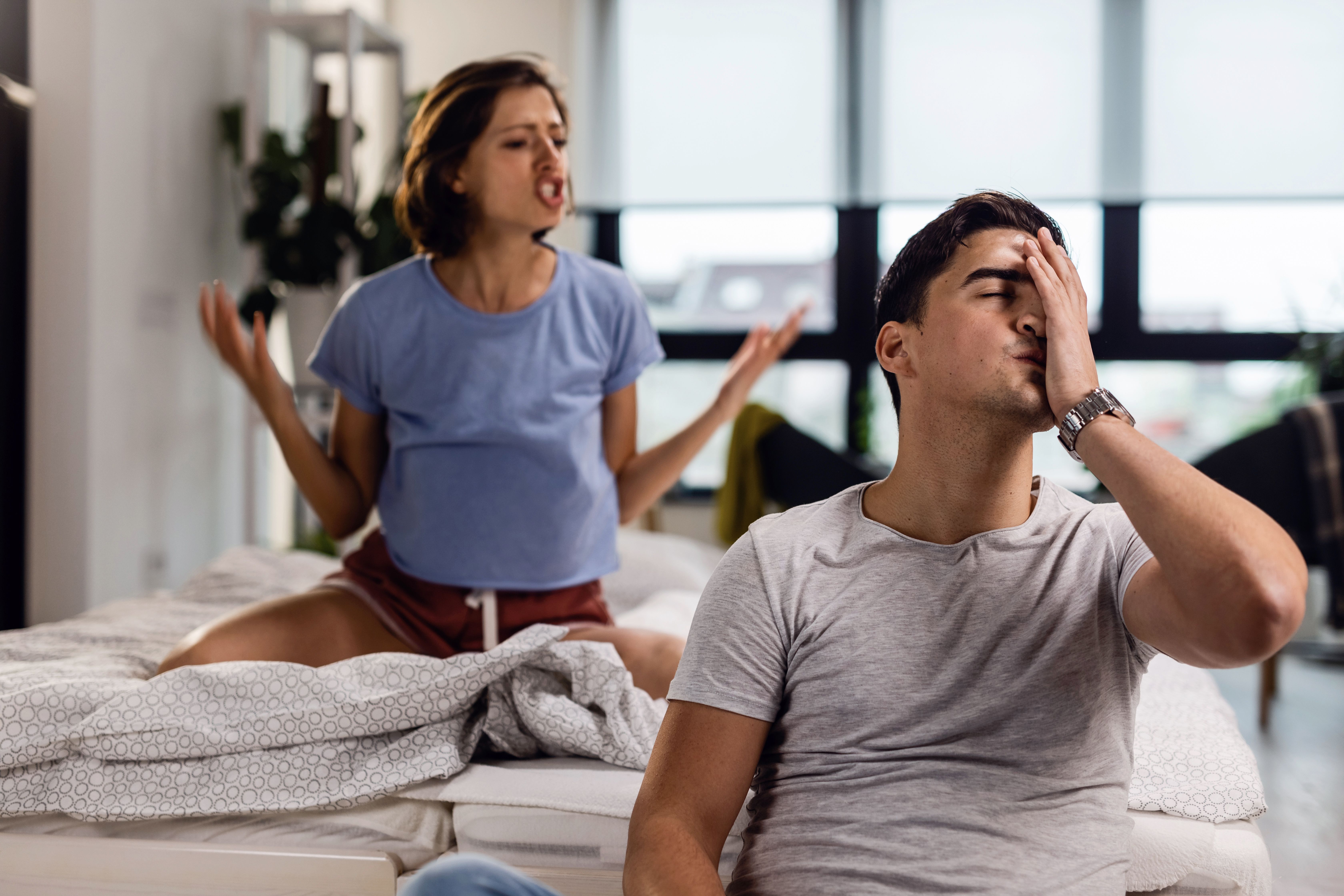 Fiatal férfi stresszes és fejfájós, miközben barátnője vitatkozik vele a hálószobában 