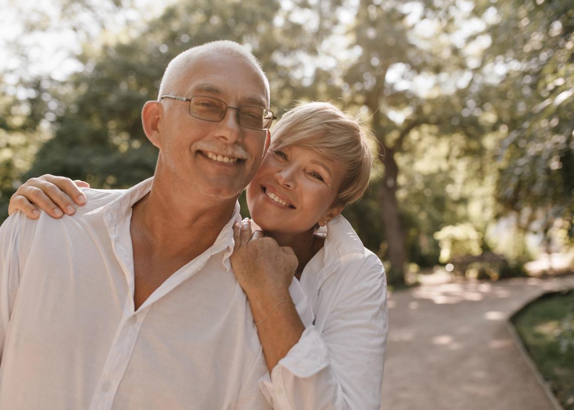 Mosolygós, ősz hajú, bajuszos, szemüveges, világos ingben lévő idős férfi szőke, fehér ruhás nővel ölelkezik a parkban.