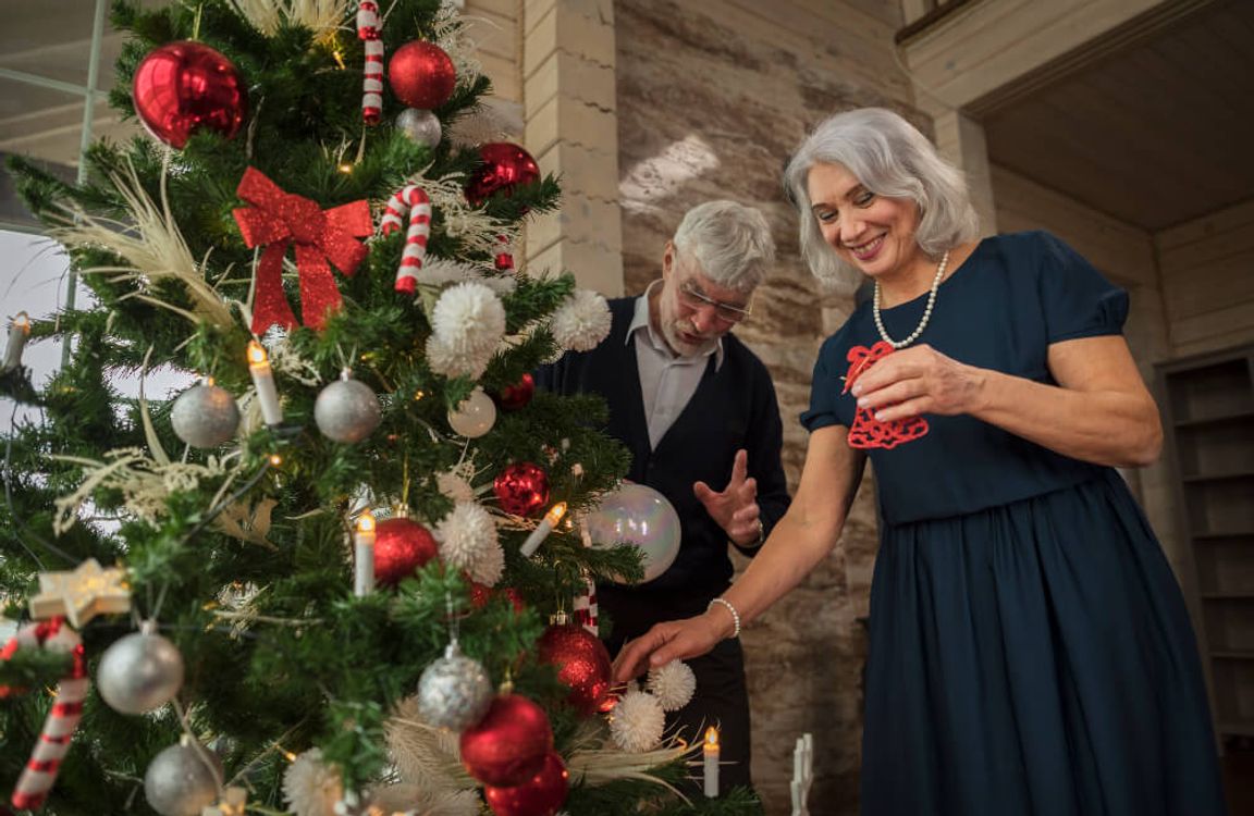 Idősebb hölgy és a a párja boldogan díszítik a karácsonyfát piros és fehér díszekkel a nappaliban, meghitt hangulatban