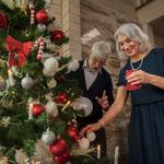 Idősebb hölgy és a a párja boldogan díszítik a karácsonyfát piros és fehér díszekkel a nappaliban, meghitt hangulatban