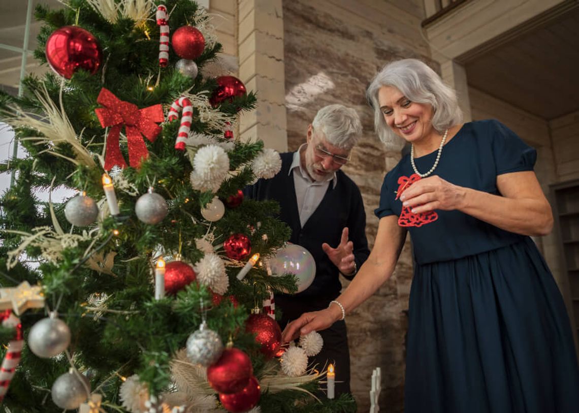 Idősebb hölgy és a a párja boldogan díszítik a karácsonyfát piros és fehér díszekkel a nappaliban, meghitt hangulatban