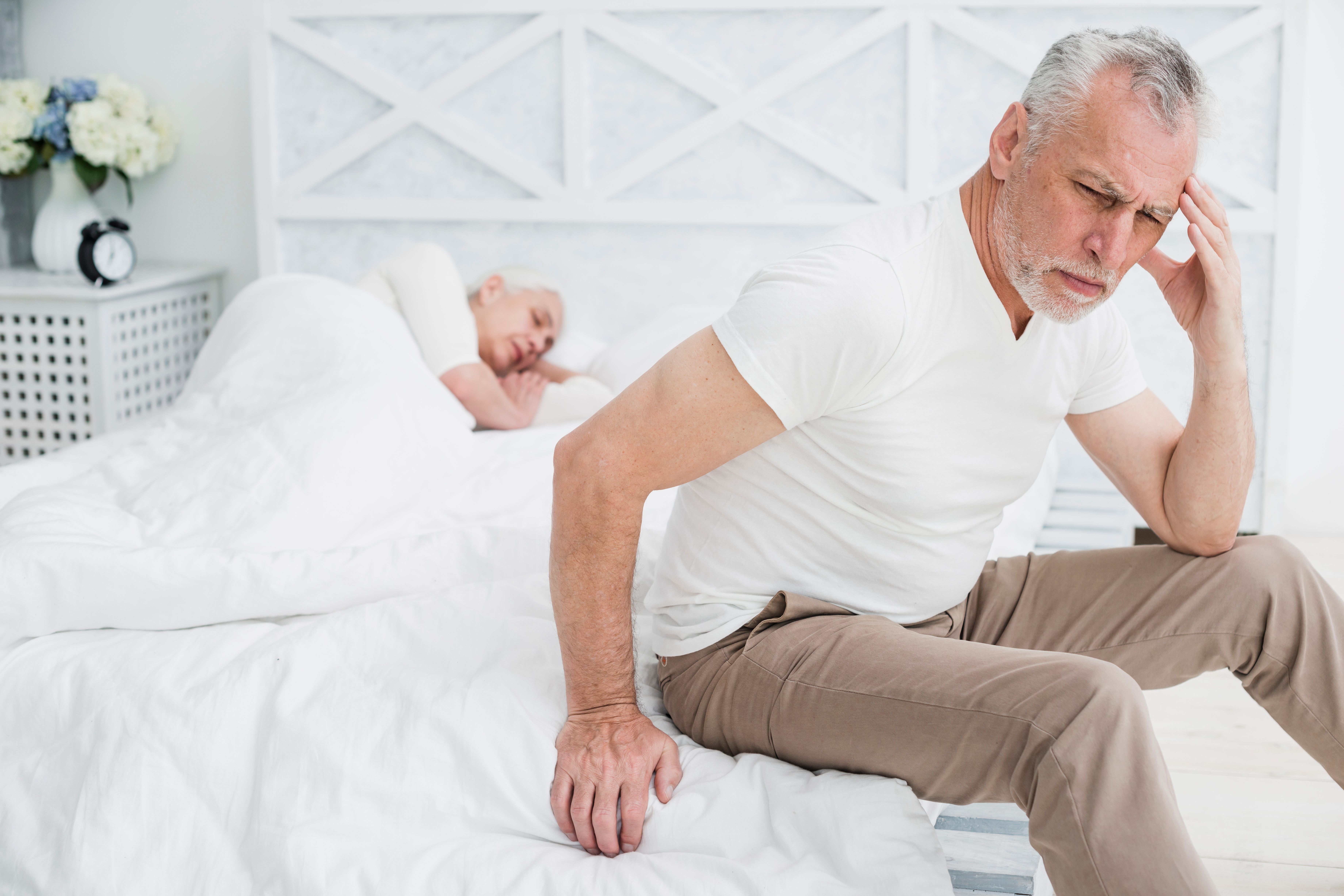 Idős férfi elgyötört arckifejezéssel ül ágya szélén, miközben a háttérben felesége még alszik