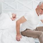 Idős férfi elgyötört arckifejezéssel ül ágya szélén, miközben a háttérben felesége még alszik