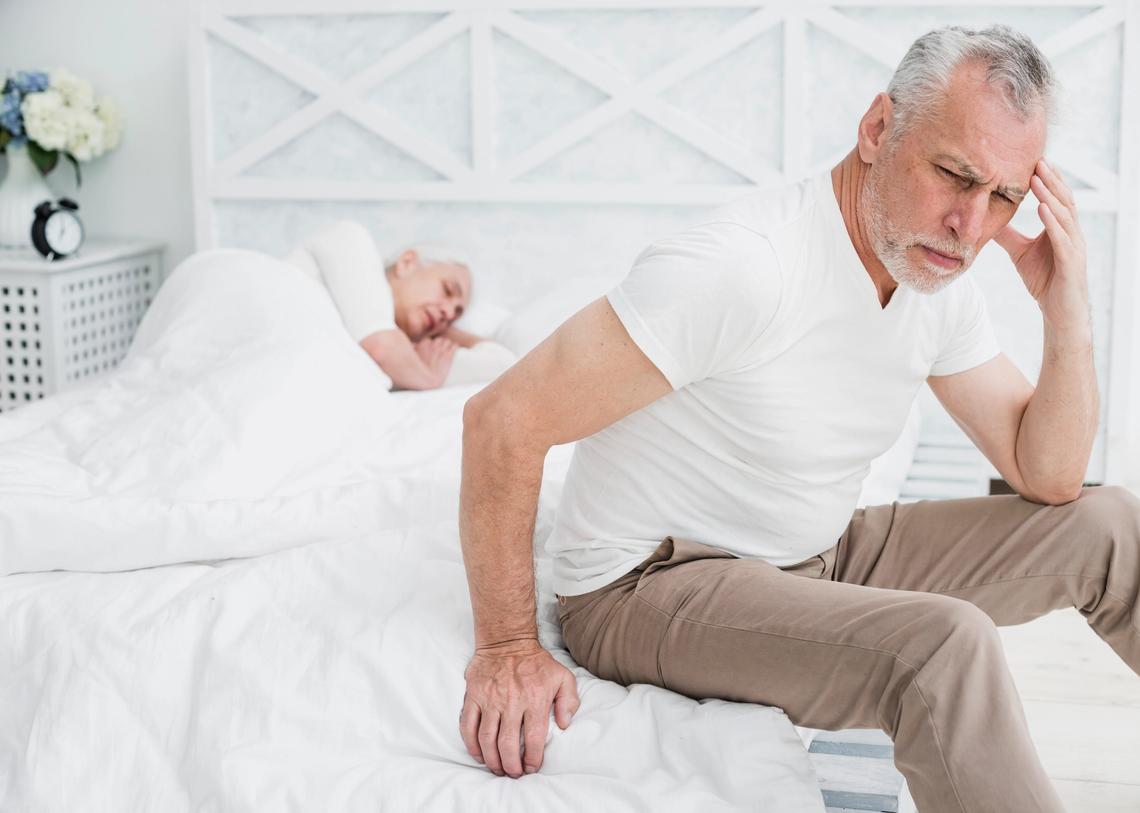 Idős férfi elgyötört arckifejezéssel ül ágya szélén, miközben a háttérben felesége még alszik