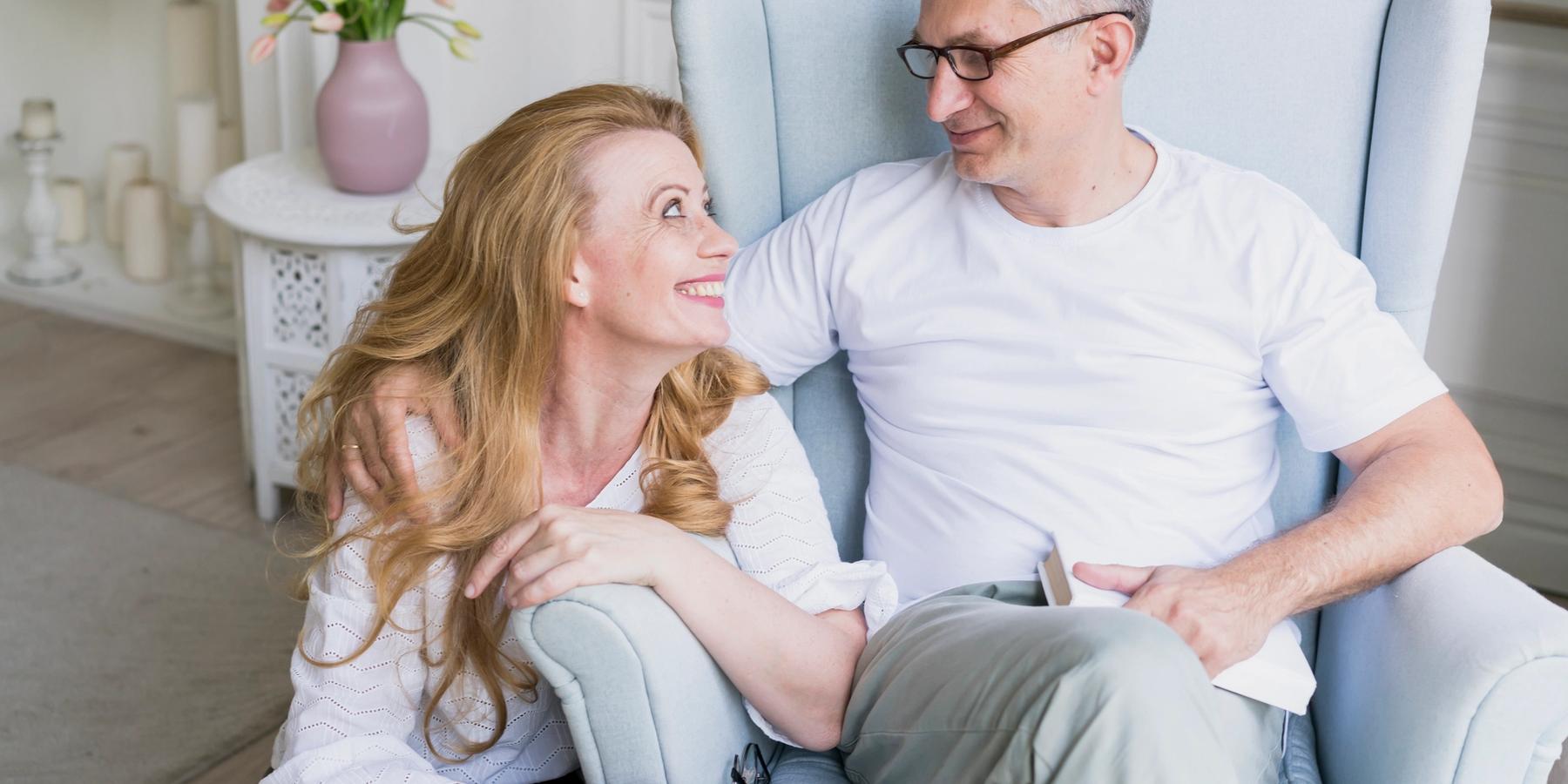 Boldog, idős pár mosolyogva néz egymásra a nappalijukban. A férfi fotelban ül, a nő mellette guggol