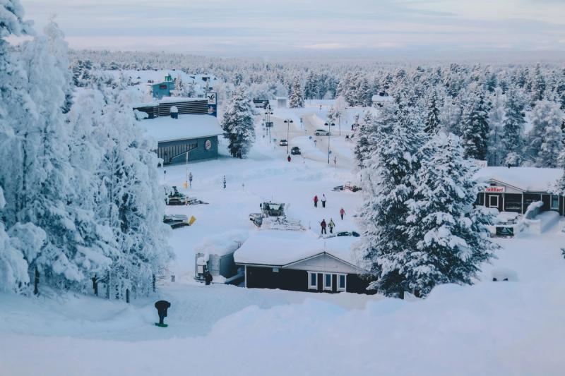 Aerial view of a snow-covered village in Espoo, Finland. The scene depicts snow-laden trees, buildings with snow-covered roofs, and people walking along snow-filled paths. The sky is overcast, suggesting a winter day.