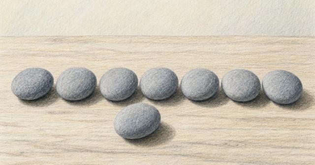 Row of seven evenly spaced smooth grey stones on a pale wood table, one nudged forward