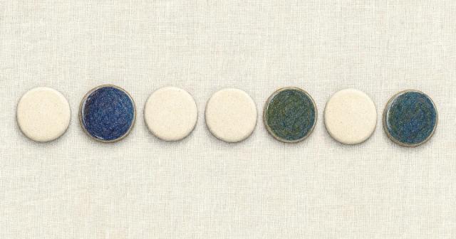 Seven small ceramic discs in a row on pale linen, three turned to a darker face