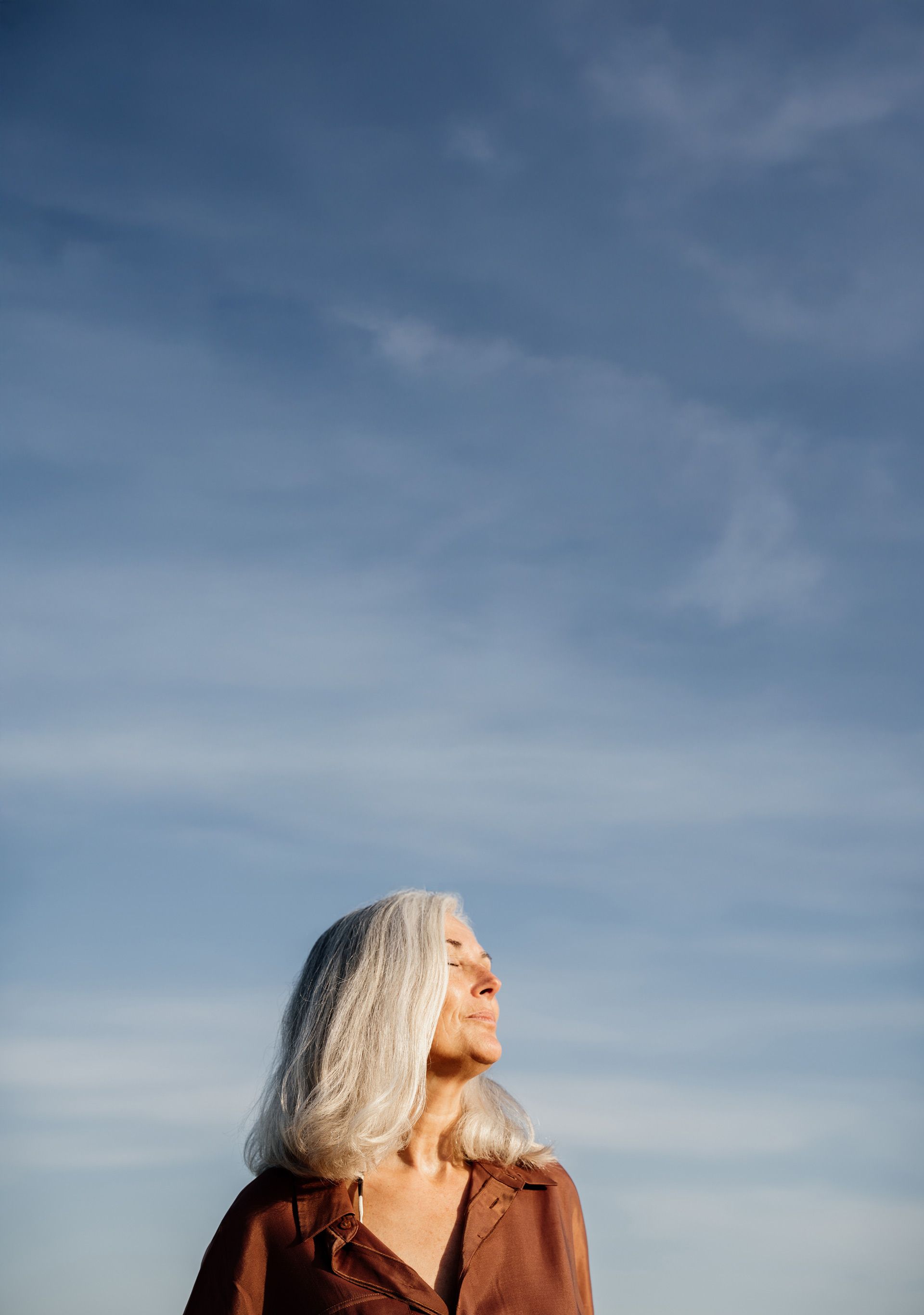 A woman in a brown shirt, looking up and basking in the afternoon sun with her eyes closed, with blue skies all around